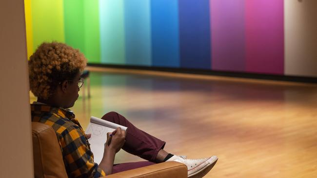 Student drawing in a sketchbook during the Lisa Marie Thalhammer exhibtion. Her colorful mural--vertical bands of colors--are in the background.